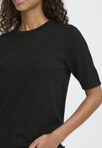 T-shirt noir à manches courtes en maille avec un col rond ; présente une texture côtelée et des poignets retroussés. Tissu lisse avec une silhouette ajustée.