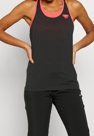 Žena v černém sportovním topu bez rukávů s korálovým lemováním a logem, a v černých sportovních kalhotách, stojící s rukama v bok.