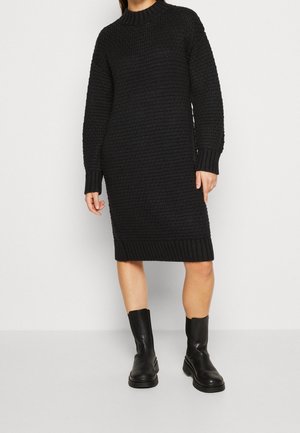 Kobieta w czarnej dzianinowej sukience swetrowej z długim rękawem i czarnych skórzanych kozakach sięgających do połowy łydki, stojąca na jasnym, jednolitym tle.