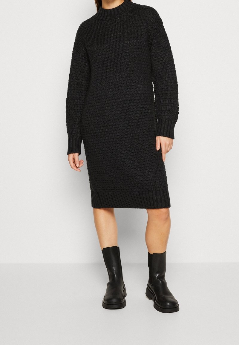 Femme portant une robe-pull noire en maille à manches longues et des bottes en cuir noires mi-mollet, debout devant un fond clair uni.
