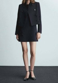 Black cropped jacket with double-breasted design, featuring large buttons. Paired with a matching black mini dress and ankle strap heels.