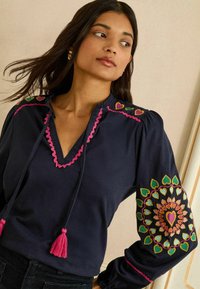 Marineblauwe blouse met roze golvende halslijn, strikjes en hartvormige borduursels op de mouwen, met kleurrijke bloemmotieven en een gestructureerde stof.