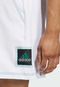 Bílý sportovní oděv s tyrkysovým prošíváním, na němž se nachází černá čtvercová nášivka s tyrkysovým a stříbrným textem "adidas EQUIPMENT".