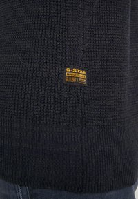 Mörkblått stickat tyg med en liten rektangulär svart och gul etikett som står "G-STAR ORIGINALS RAW DENIM" sydd nära nederkanten.