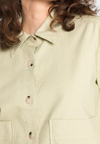 Ljusgrön texturerad skjorta med krage, fyra knappar och två framfickor. Tyget har ett subtilt rutmönster.