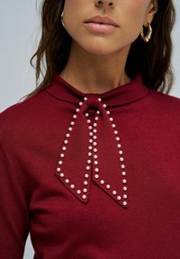 Mujer con una blusa roja adornada con un lazo decorativo contorneado de cuentas de perlas cerca del escote y aretes de aro dorados visibles.