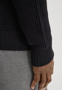 Pull en maille noire avec un motif texturé, des poignets et un ourlet côtelés. Main reposant sur un fond en tissu gris.