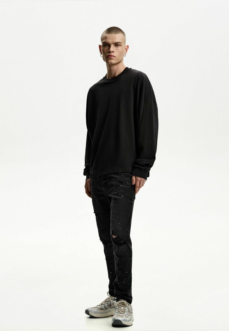 Cămașă neagră oversized cu mâneci lungi, blugi negri distruși cu stropi de vopsea și adidași gri cu accente bej.