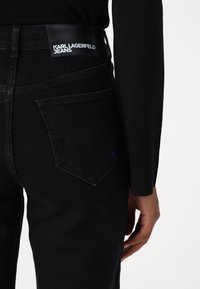 Schwarze Denim-Jeans im Slim Fit, mit einem markanten Markenlogo-Patch an der Taille und einem dezenten blauen Akzent auf der Gesäßtasche.