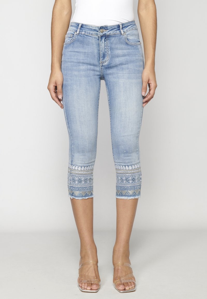 Jean en denim bleu clair raccourci avec broderie décorative près des ourlets, porté par une personne debout avec des sandales à brides transparentes.
