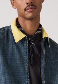 Άνδρας που φοράει τζιν μπουφάν με κίτρινο γιακά και μαύρο μπαντάνα δεμένο γύρω από τον λαιμό πάνω από μαύρο πουκάμισο.