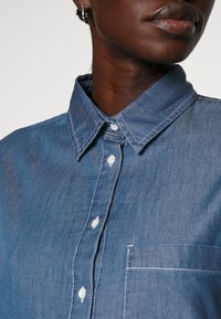 Spijkershirt in een mediumblauwe tint met een gestructureerde kraag, knopen aan de voorkant, witte stikselaccenten en een borstzak. Soepele textuur.