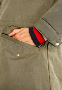 Chaqueta verde oliva con una mano en el bolsillo lateral. Costuras visibles, botón a presión dorado y un detalle de puño en rojo, azul marino y blanco que contrasta.