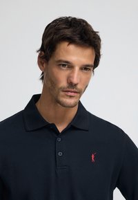 Svart pikétröja i texturerat tyg med en treknappslå och en liten röd broderad logotyp på vänster bröst.