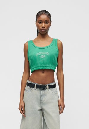 Femeie cu părul împletit, purtând un top verde scurt Quiksilver și blugi gri deschis cu talie înaltă și o curea neagră.