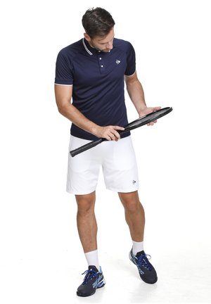 Marineblauw poloshirt met witte accenten, witte sportshorts en donkere tennisschoenen. Houdt een zwarte tennisracket vast terwijl hij naar beneden kijkt.