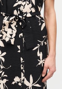 Gros plan sur une robe noire à imprimé floral crème, avec boutons sur le devant et ceinture nouée à la taille, montrant une partie du bras et de la main d'une personne.