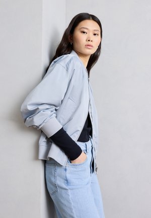 Mlajša ženska z dolgimi temnimi lasmi, oblečena v svetlo modro bomber jakno, črno majico z dolgimi rokavi in svetlo modre kavbojke, stoji ob sivi steni.