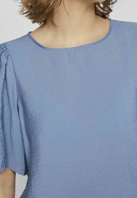 Blouse bleu clair en tissu doux et texturé, avec des manches courtes bouffantes et un col rond. Design simple avec des détails minimaux.