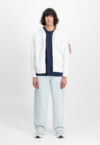 Sudadera blanca con cremallera y camiseta navy debajo, jeans de pierna ancha de color azul claro y zapatillas grises. La sudadera tiene un detalle de etiqueta en el bolsillo.