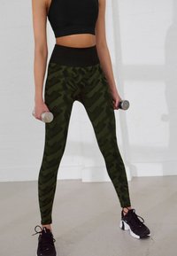 Mustat cropped topit yhdistettynä korkeavyötäröisiin vihreisiin maastokuvioisiin legginsseihin. Legginsseissä on sileä pinta ja kapeahko muotoilu. Kädessä käsipainot.