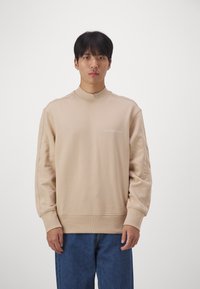 Felpa beige con collo alto, dotata di un piccolo logo "Calvin Klein Jeans" in bianco. Tessuto morbido e vestibilità ampia, abbinata a jeans blu.