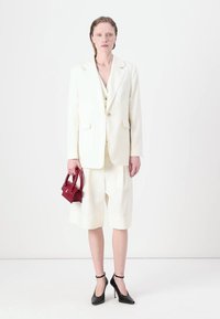 Weißer, übergroßer Blazer kombiniert mit einer cremfarbenen Weste und knielangen Shorts. Accessoires umfassen eine kleine rote Handtasche und schwarze High Heels.