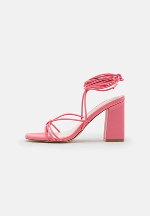 Pink high-heeled sandal with a block heel, featuring thin crossed straps and a knotted ankle tie; smooth surface and open toe design.