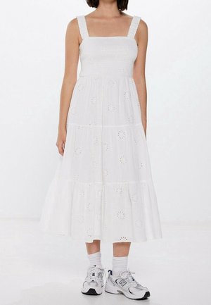 Frau trägt ein weißes ärmelloses Midikleid mit floralen Ausschnitten, kombiniert mit weißen Socken und weißen Sportschuhen vor einem schlichten Hintergrund.