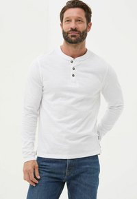 Weißes Langarm-Henley-Shirt mit drei schwarzen Knöpfen am Ausschnitt, strukturierter Stoff, kombiniert mit blauen Jeans, Frontansicht.