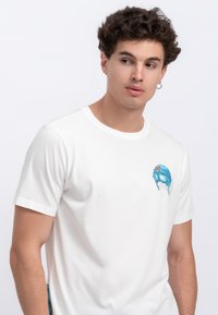 T-shirt bianco a maniche corte realizzato in cotone morbido, con una grafica blu di un casco con occhiali sul lato sinistro del petto.