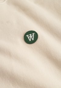 Cream-colored fabric featuring a green circular embroidered patch with a white "W" and "A." The texture appears smooth and knit.