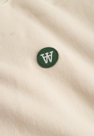 Cremefarbener Stoff mit einem grünen, kreisförmigen gestickten Patch, auf dem ein weißes "W" und "A" zu sehen sind. Die Textur wirkt glatt und gestrickt.