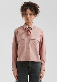 Chemise à rayures rose clair avec deux poches poitrine, manches longues et une écharpe florale au cou. Tissu doux et tissé avec une coupe décontractée.