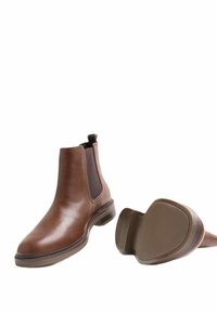 Hnědé kožené Chelsea boty s elastickými stranami, plochou gumovou podrážkou, zaoblenou špičkou a poutkem na zadní straně. Jedna bota leží na boku.