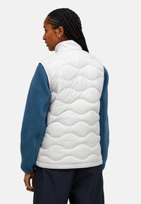 Vadderad väst med en vit, vågmönstrad front och ärmar i blå fleece. Hög krage och avslappnad passform. Lätt material.