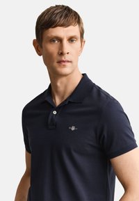 Mann mit kurzen braunen Haaren, der ein marineblaues Polohemd mit kleinem gesticktem Logo auf der Brust trägt und leicht seitlich schaut.