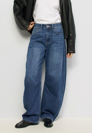 Persona che indossa jeans blu a gamba larga, t-shirt bianca, giacca di pelle nera e scarpe nere, in piedi contro un muro semplice.