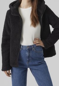 Svart zip-up fleecejacka som bärs öppen över en vit ribbad långärmad topp, kombinerad med höga blå jeans, hand på höften.