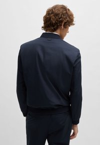 Veste de bombardier marine en tissu léger, avec un col et des poignets côtelés, un design épuré et court, et un branding minimal.