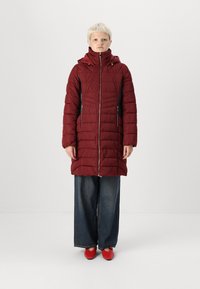 Manteau rembourré bordeaux avec un col haut, des zips latéraux et une couture texturée, associé à un jean évasé et des chaussures rouges.