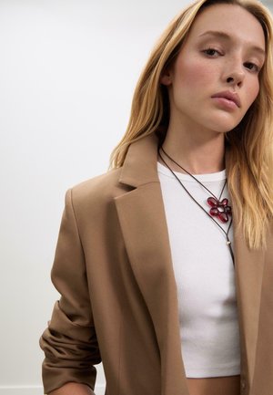 Brauner taillierter Blazer über einem weißen gerippten Crop-Top, ergänzt durch eine rote Blumenanhänger-Halskette an einem dunklen Band. Glatte Textur, taillierter Schnitt.