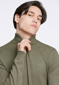 Suéter verde oliva con cuello alto y detalle de cremallera, hecho de un suave tejido de punto, con puños acanalados y un diseño ajustado.