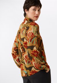 Blusa a fantasia floreale con maniche lunghe, caratterizzata da colori ricchi come oro, rosso e turchese. Tessuto morbido con vestibilità rilassata e orlo arrotondato.
