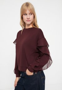 Νεαρή γυναίκα φοράει ένα βυσσινί μπλουζάκι με μακριά μανίκια και διαφάνειες, καθώς και σκούρα μπλε τζιν, στέκεται μπροστά σε έναν απλό φωτεινό φόντο.