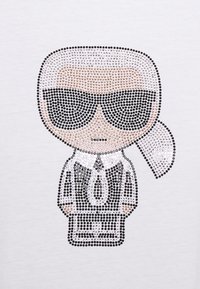 Gráfico adornado con piedras de strass que presenta un personaje con gafas de sol negras, un cuello blanco y un traje negro sobre un fondo blanco.