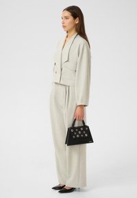 Donna in un completo beige su misura, che tiene in mano una borsa nera con borchie argentate, indossando tacchi a punta neri, in piedi contro uno sfondo neutro.