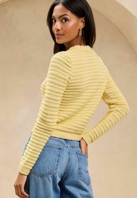 Dzeltena adīta džemperis ar teksturētām svītrām un viļņotu dekoltē, saskaņots ar ziliem augstvērtīgiem džinsiem, demonstrējot brīvu stilu.