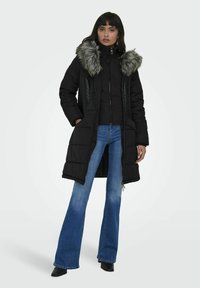 Svart pufferjacka med fuskpäls på huven, dragkedjestängning, sidofickor och kviltad textur, i kombination med utställda blå jeans och svarta stövlar.