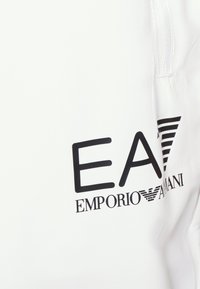 Baltas audinys, ant kurio pavaizduotas didelis juodas logotipas "EA7 EMPORIO ARMANI" su horizontaliomis juostomis, akcentuojantis modernų dizainą ir prekių ženklą.